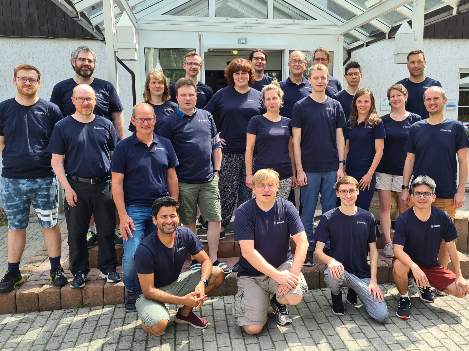 Group photo of the BENOCS team, wearing matching navy blue shirts, standing and sitting outside a building.